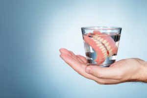 Hand holding a glass of water with dentures in it in front of a gray background
