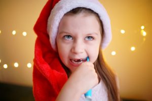 Little girl in a Santa hat brushing her teeth in front of holiday lights